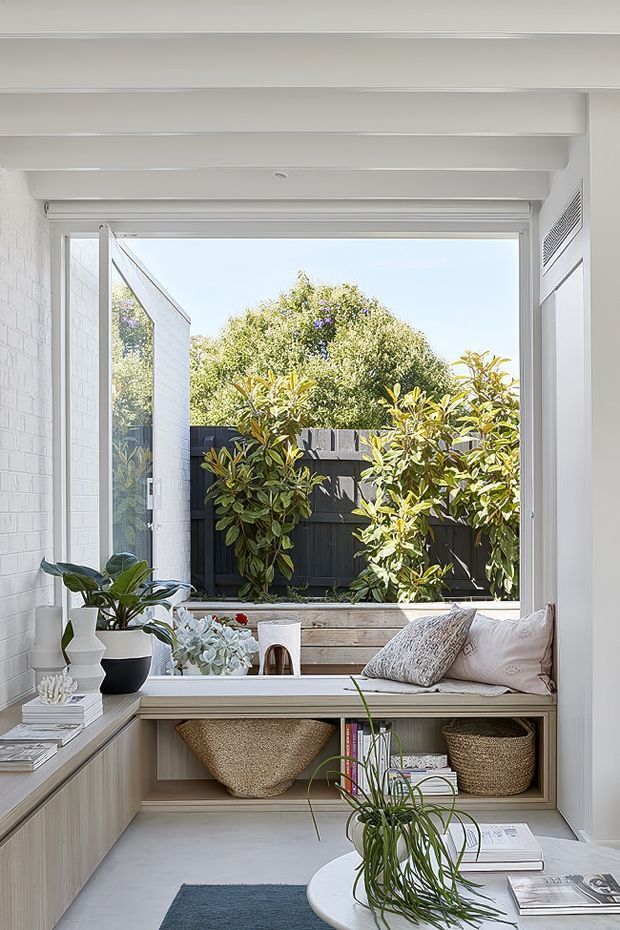 Bright, compact interior nook centred on a large open window overlooking a planted courtyard. The space features a built-in window seat with cushions, pale timber cabinetry and open shelving with woven baskets, white painted brick walls, exposed white ceiling beams, potted plants, and a small round table with magazines in the foreground. The overall look is light, natural, and relaxed.