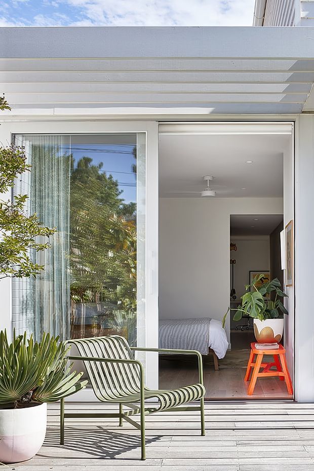 Bright view from a timber-clad outdoor deck into a compact bedroom through an open sliding glass door. The white exterior has a projecting louvred or slatted canopy, while the room contains a bed, ceiling fan, indoor plant and vivid orange-red stool. A striped outdoor lounge chair and potted plants occupy the foreground, with trees reflected in the glazing.