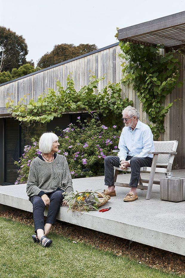 An outdoor courtyard or terrace beside a modern single-storey building. A raised concrete patio with built-in seating steps is surrounded by climbing vines and flowering plants. Two older adults sit outdoors, with a lightweight outdoor chair beneath a timber pergola. The building has a long weathered vertical timber-clad wall, dark-framed glazing, and a simple low-sloping roofline. The scene is bright, relaxed, and garden-integrated.