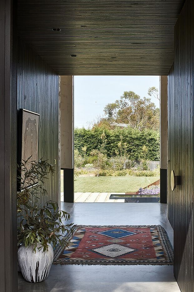 A dark, minimalist entry passage framed by deeply textured vertical wall and ceiling cladding, opening onto a landscaped garden and reflecting pool. The polished grey concrete floor, recessed ceiling lights, sculptural wall artwork, potted plant and patterned runner create a dramatic transition between interior and exterior.