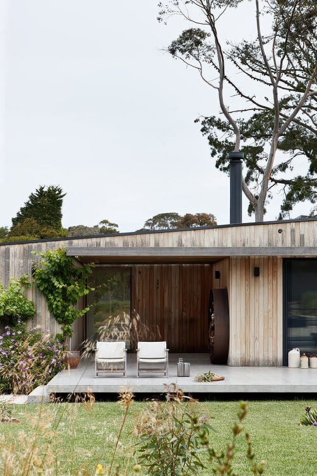 Front exterior view of a low-slung contemporary house set within a landscaped garden. The facade is clad in vertically arranged weathered timber, with a broad flat roof, dark-framed glazing, a recessed covered entry, and a slim raised concrete terrace. Outdoor lounge chairs sit on the terrace beside climbing greenery and flowering planting. A dark cylindrical chimney or flue rises above the roofline.