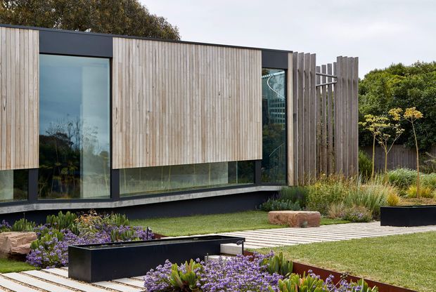 Contemporary low-profile building exterior with extensive vertical timber cladding, dark framing, and large floor-to-ceiling glazing. The landscaped foreground includes lawn, raised black planters, ornamental grasses, purple flowering plants, timber paving, and a sculptural stone element. A vertical timber-screened section creates a partially enclosed architectural feature at the right.
