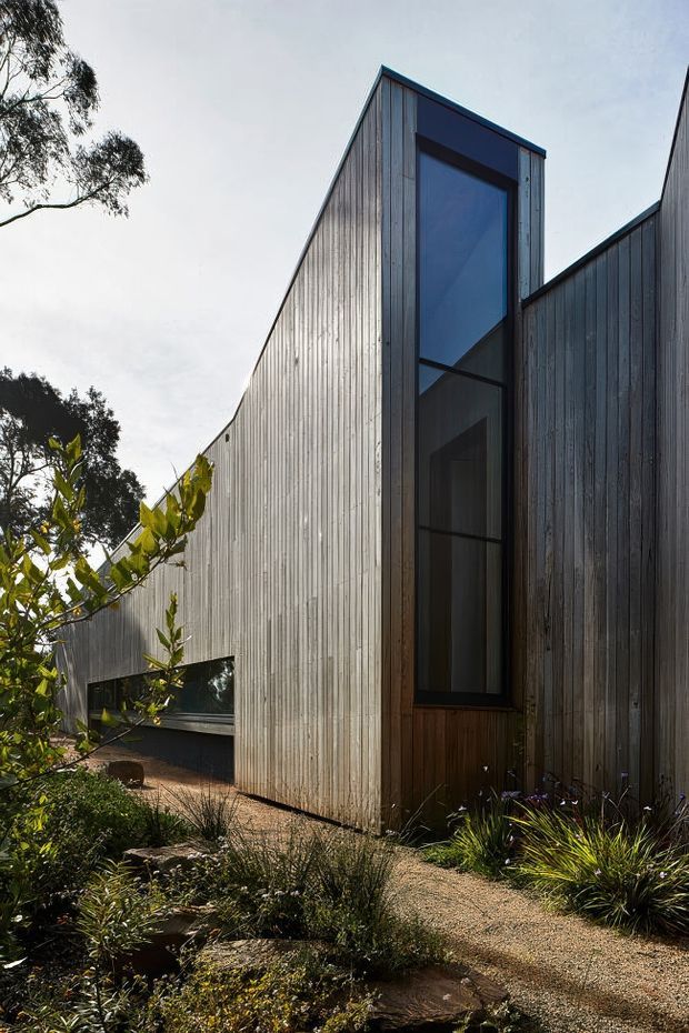 Exterior view of a contemporary, sharply geometric building set within landscaped gardens. The facade is clad in vertically ribbed, weathered grey timber, with a tall narrow glazed opening framed in dark timber or metal. Angular roof and wall forms create a strong sculptural silhouette, while a gravel garden path and native planting lead toward the entrance.