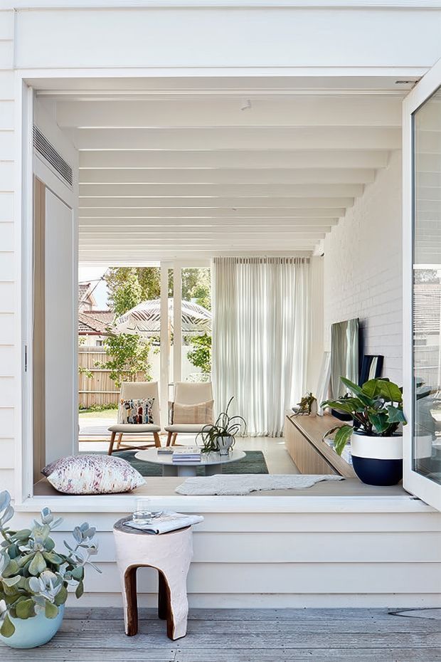 Bright coastal-style living room viewed from an exterior deck through a large open window. The room features white-painted exposed ceiling beams, white walls, sheer curtains, a low timber media console, two lounge chairs, a coffee table, and indoor plants. A covered outdoor or semi-enclosed terrace extends toward the rear garden, with strong indoor-outdoor connection and abundant natural light. The exterior framing and decking are also visible in the foreground.