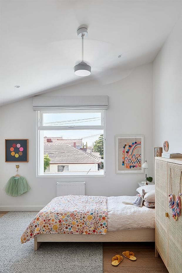 A bright, compact bedroom with white walls and a sloped ceiling, centered on a window overlooking neighboring rooftops. The room contains a low timber bed with white bedding and a colorful floral-patterned quilt, a woven-texture wardrobe, patterned artwork, playful wall decor, a ceiling pendant, and a soft grey carpet. The overall look is cheerful, airy, and eclectic with natural woven textures and colourful accents.