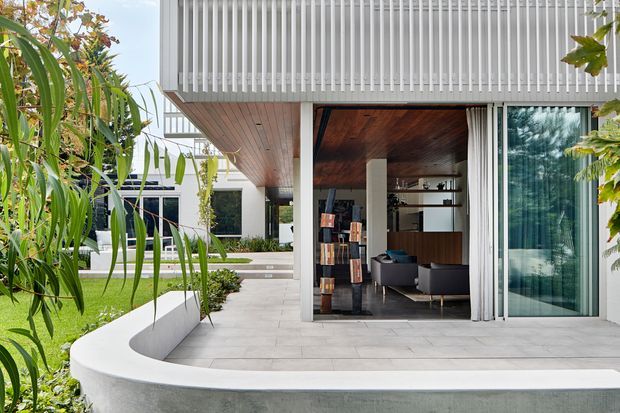 A contemporary residential courtyard and covered outdoor living area. The image shows a white rendered curved planter or boundary wall, pale tiled paving, lush planting, and a sheltered sitting room opening through large glazed sliding doors. The building has a strong horizontal composition, white vertical screening or fins above, a timber-lined soffit, and dark timber interior finishes visible through the opening.