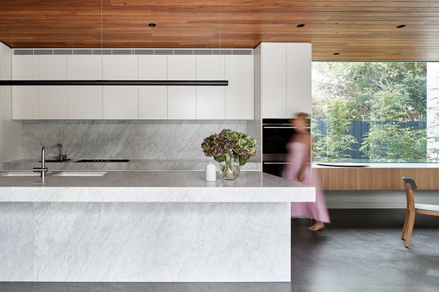 Contemporary kitchen with a large waterfall island in pale veined stone, white handleless cabinetry, a matching stone backsplash, and warm timber lining across the ceiling and rear cabinetry. A dark linear pendant runs above the island, while a large window brings in views of greenery and daylight. An adjacent dining area with a timber table and chair is visible beyond the kitchen.
