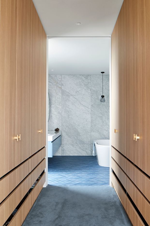 A narrow, refined dressing corridor with floor-to-ceiling warm timber wardrobe cabinetry and integrated drawers on both sides, opening directly into a contemporary bathroom. The bathroom features pale grey marble-look wall panels, a blue-grey herringbone tile floor, a white freestanding bathtub, a pale vanity, and a black pendant light. A grey carpet runs through the wardrobe area, with brass-gold handles providing a small accent.
