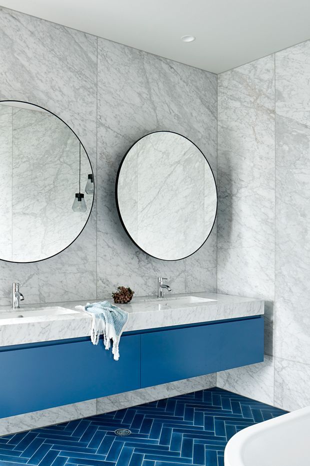 A contemporary bathroom featuring full-height white veined marble wall cladding, a matching marble double vanity benchtop, vivid blue floating cabinetry, and a deep blue herringbone tile floor. Two large round mirrors with slim black frames sit above the basins, accompanied by minimalist pendant lights. The corner of a white freestanding bathtub is visible in the foreground.