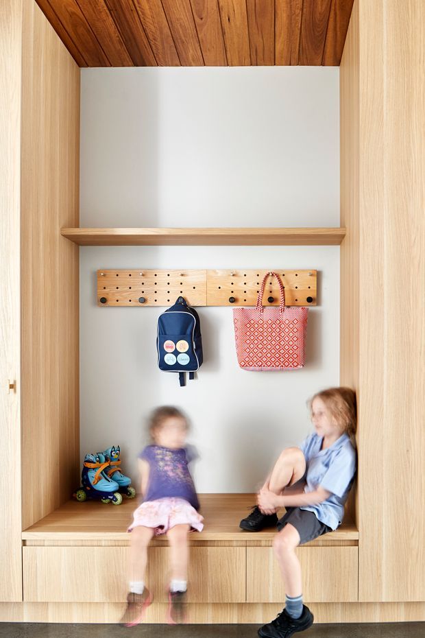 A built-in timber entryway mudroom with full-height light wood cabinetry framing an open alcove. The alcove has a white painted back wall, a warm timber ceiling, a shelf, pegboard hooks with children's bags, a bench seat with integrated drawers, and roller skates. Two children sit in the built-in bench area. The space is bright, warm, and highly functional.
