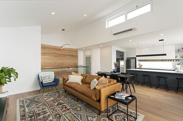 Bright open-plan living and kitchen interior with a warm tan sectional sofa, patterned area rug, black coffee table, and a compact blue armchair. The kitchen features a long dark island with integrated seating, white cabinetry, pendant lighting, and timber-look flooring. A large horizontal timber feature wall adds warmth, while high clerestory windows and recessed lighting create an airy contemporary space.