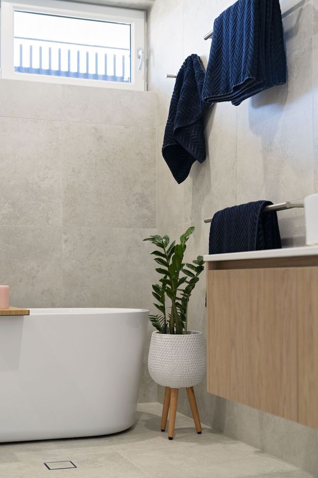 A bright, contemporary bathroom featuring a freestanding white bathtub, large pale stone-look wall and floor tiles, a timber-toned floating vanity, chrome towel rails with navy towels, and a potted indoor plant. Natural light enters through a high horizontal window.