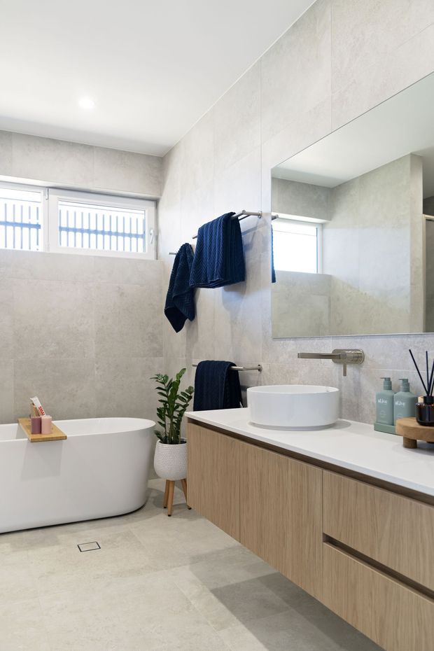 Bright contemporary bathroom featuring a freestanding white bathtub, wall-mounted timber-look vanity with a white benchtop, vessel basin, large frameless mirror, pale stone-look wall and floor tiles, and brushed metal tapware. A high horizontal window provides natural light, while dark navy towels add contrast.