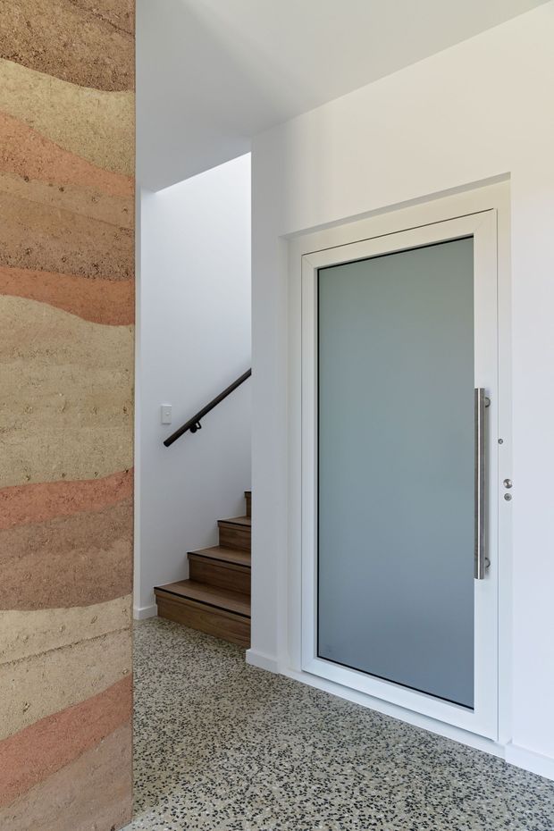 A bright contemporary entryway featuring a large frosted-glass front door with a white frame and vertical metal handle, adjacent to a stairway with dark timber treads and a simple dark handrail. The space has polished terrazzo flooring and a textured, horizontally banded feature wall in muted beige and terracotta tones.