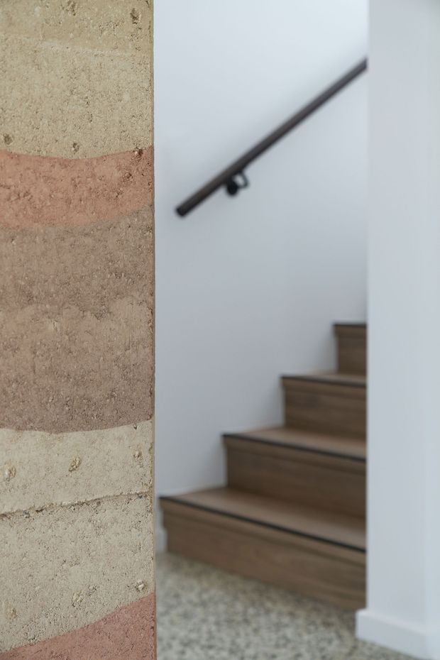 Interior architectural detail showing a timber staircase ascending beside a white wall with a dark slim handrail. A textured exposed brick wall with alternating pale and muted terracotta bands frames the left side, while a speckled hard floor and white skirting are visible below.