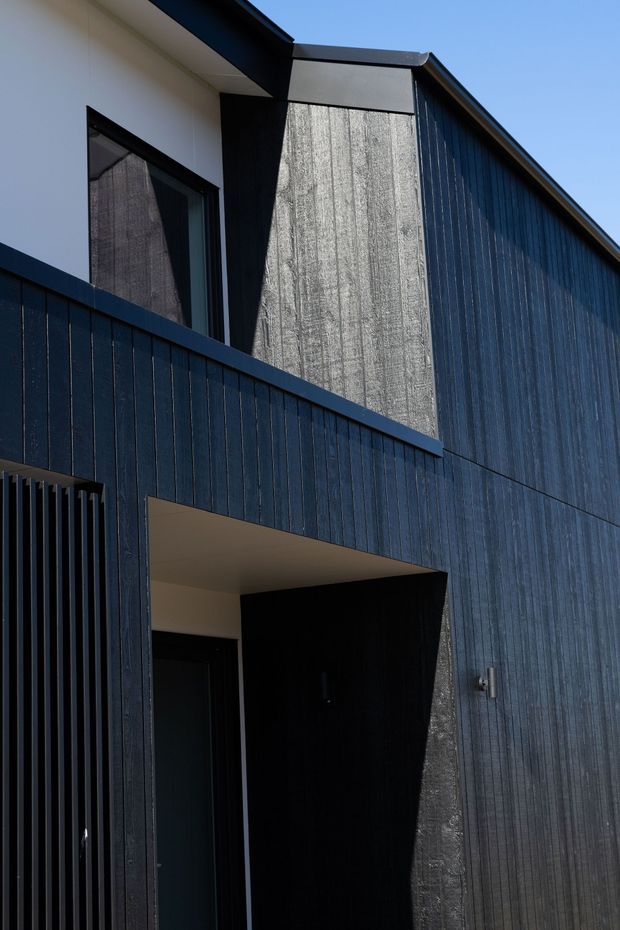 Close-up exterior view of a contemporary multi-level building with dark vertically textured cladding, a sharply angled gabled roofline, contrasting white rendered surfaces, black-framed glazing, and a recessed entry or passage beneath an upper balcony. The composition emphasizes strong geometric forms, deep shadows, and crisp modern detailing.