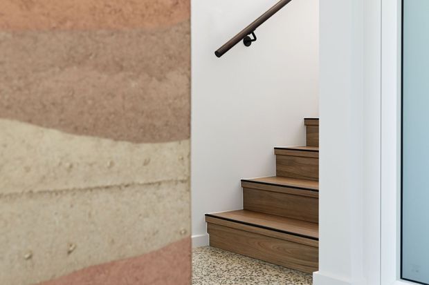 A close-up interior view of a contemporary stairway with dark timber treads and risers, a simple dark metal handrail, white walls, and a terrazzo-like speckled floor. A layered textured wall surface in muted earthy tones forms the foreground, while a bright glazed opening is visible at right.