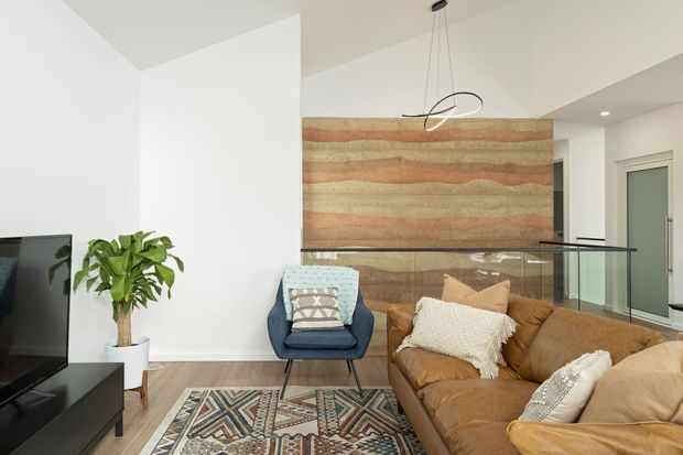 Bright contemporary living room with a large tan leather sectional, patterned area rug, blue accent chair, indoor plant, and a sculptural pendant light. A striking horizontal timber feature wall forms the backdrop, with a glass balustrade revealing a stair or landing beyond.