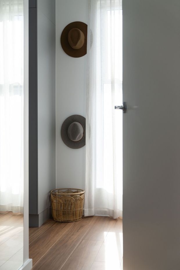 A softly lit interior corner viewed through a partially open door, featuring two wall-mounted hats, sheer white curtains over tall glazing, warm timber flooring, and a woven rattan basket. The space has a simple, airy, natural aesthetic with strong daylight and minimal furnishings.