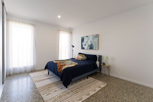 Bright, simply styled bedroom with a navy upholstered bed, patterned bedding, a neutral geometric rug, pale walls, sheer floor-to-ceiling curtains, a small timber side table, floor lamp, and abstract artwork. The room has broad windows and a speckled hard floor, creating an airy contemporary feel.