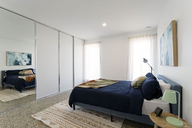 Bright contemporary bedroom with a navy upholstered bed and bedding, mirrored floor-to-ceiling wardrobe doors, light carpet, layered patterned rugs, sheer curtains, and pale walls. A small timber bedside table with a mint-green lamp and framed artwork add restrained colour accents.