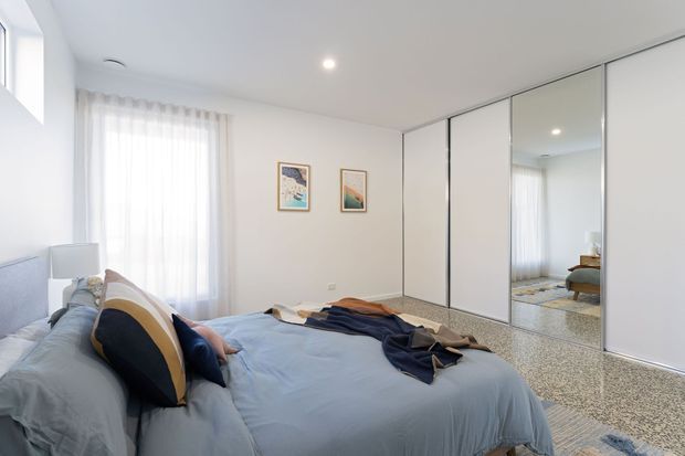 Bright, contemporary bedroom with a low-profile bed dressed in pale blue bedding and layered navy, cream and mustard cushions. Full-height mirrored wardrobe doors line one wall, reflecting the bed and window. The room has white walls and ceiling, sheer curtains, recessed lighting, small framed artworks, and a speckled carpeted floor.