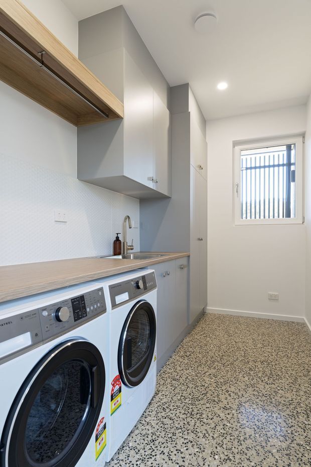 Bright, narrow laundry room with front-loading washer and dryer beneath a wood-look benchtop. The room includes pale grey and white cabinetry, an open timber shelf with hanging rail, a small sink with mixer tap, subtle white wall tile backsplash, and a terrazzo-style floor. A barred window provides natural light.