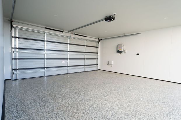 Bright, spacious modern residential garage with a sectional automatic garage door, smooth white walls and ceiling, ceiling-mounted door opener, wall-mounted electrical equipment, and a speckled epoxy-style floor coating.