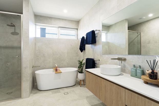 Bright contemporary bathroom featuring a freestanding white bathtub, frameless glass shower, wall-to-wall pale stone-look finishes, a timber-look floating vanity with a white countertop and vessel basin, large mirror, and towel rails. Natural light enters through a high horizontal window.