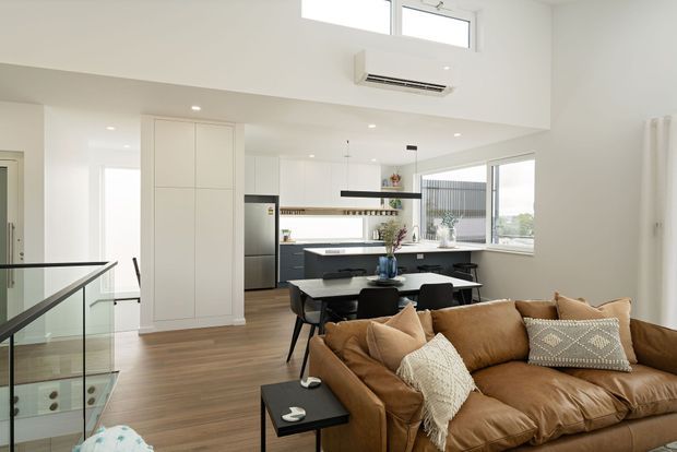 Bright open-plan contemporary interior combining a living room, dining area and kitchen. The space features a large tan leather sectional sofa, black dining furniture, white built-in cabinetry, a dark kitchen island, timber flooring, expansive windows, and a stairwell with a glass balustrade. The high ceiling and upper clerestory windows create an airy, spacious feel.