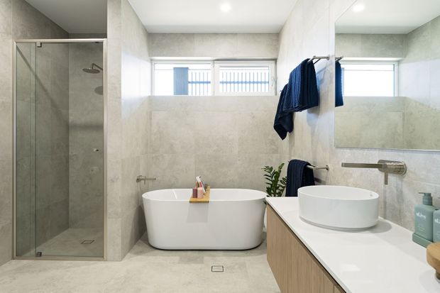 Bright contemporary bathroom featuring a freestanding white bathtub, frameless glass shower enclosure, wall-mounted mirror, floating timber vanity with a white vessel basin, and large-format pale grey-beige stone-look wall and floor finishes. Natural light enters through high horizontal windows.