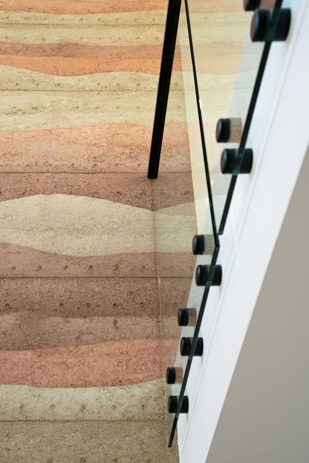 Close-up architectural detail of an interior stairway with a clear glass balustrade secured by black circular metal fittings. The stairs and adjacent wall feature a textured, horizontally banded finish in muted cream, beige, terracotta, and brown tones. A slim black stair or handrail element contrasts with the pale surfaces.