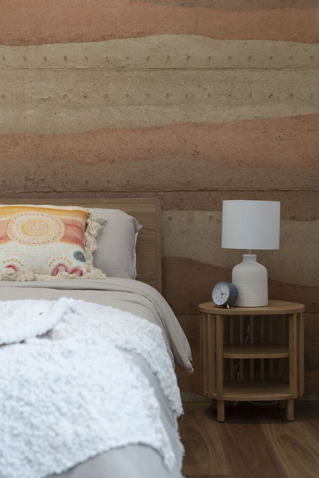 A close-up bedroom interior featuring a bed with neutral grey bedding, a colorful patterned fringed cushion, and a soft white throw. A round wooden bedside table with open shelving holds a white ceramic lamp and small clock. The room has a warm, earthy textured earthen wall finish and wood-look flooring, creating a cozy natural atmosphere.