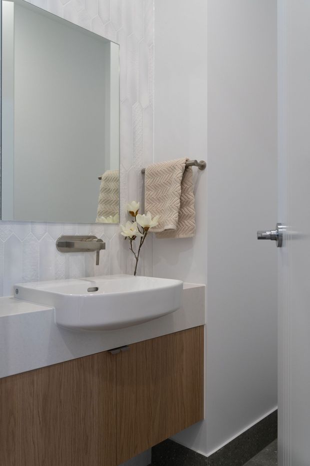 A compact contemporary bathroom featuring a wall-mounted white basin on a white benchtop, warm wood-grain vanity cabinetry, a large frameless mirror, brushed metal wall-mounted tapware, and textured white geometric wall tiles. A neutral woven hand towel and small floral branch add softness.