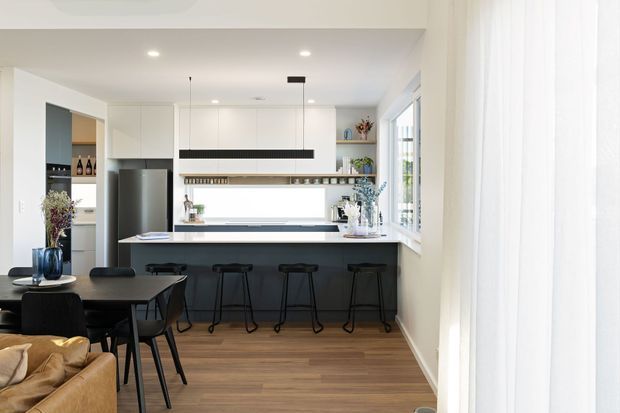 Bright contemporary open-plan kitchen and dining area with a dark island peninsula, four black bar stools, white cabinetry, integrated stainless-steel refrigerator, timber flooring, and linear pendant lighting. A dining table and part of a living area are visible in the foreground, while sheer curtains cover large windows on the right.