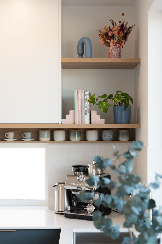 A bright, carefully styled kitchen coffee station with open timber shelving, white cabinetry, a built-in niche, coffee machine and accessories. The shelves display cups, jars, books, a potted plant, decorative dried flowers and a sculptural blue object. The palette is predominantly white and light timber with muted blue-green accents.