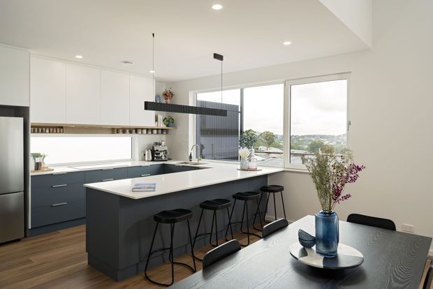 A bright contemporary open-plan kitchen and dining area with white full-height cabinetry, dark blue-grey lower cabinets, a large white island with seating for four, and integrated appliances. A long black linear pendant hangs above the island, while a dark dining table with a blue vase occupies the foreground. Large windows provide views to the leafy suburban surroundings.
