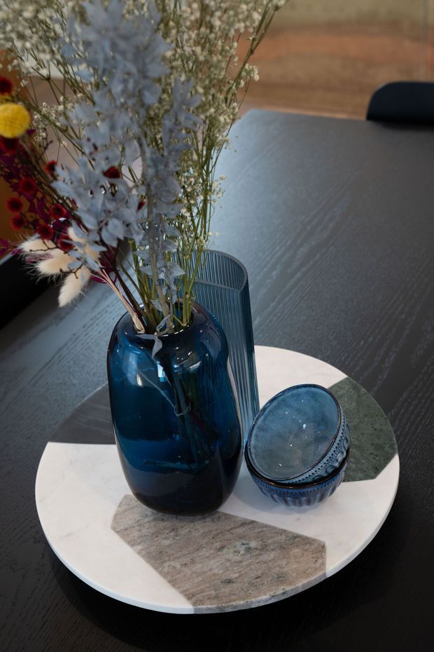 Close-up still life of a dark dining or coffee table featuring a large glossy deep-blue glass vase filled with dried flowers, a smaller matching blue glass vessel, and a round white stone or marble tray with contrasting grey-brown inlays. A dark upholstered chair and warm timber-toned background are partially visible.
