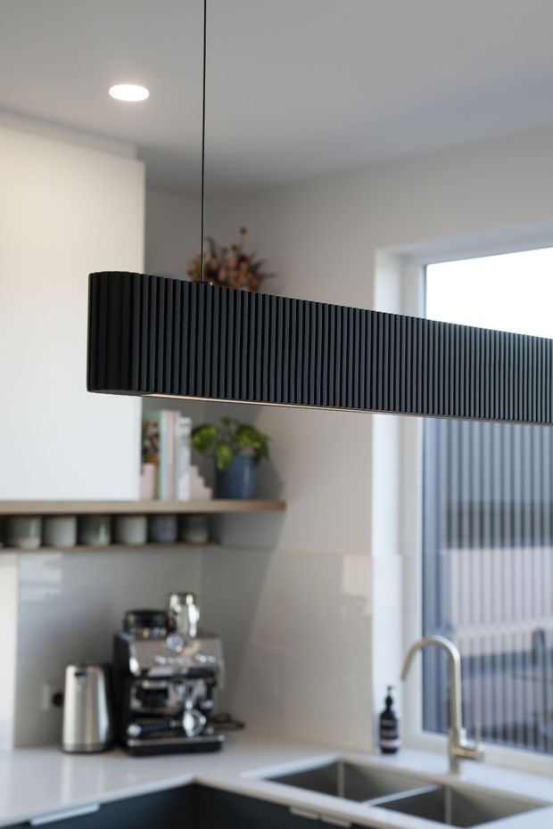 A close-up view of a contemporary kitchen featuring a long black vertically ribbed pendant light suspended over a pale stone or composite benchtop. The kitchen includes dark lower cabinetry, glossy light backsplash panels, open timber shelving with ceramics and plants, a coffee machine, stainless-steel appliances, and a double-bowl sink beside a window with vertical blinds.