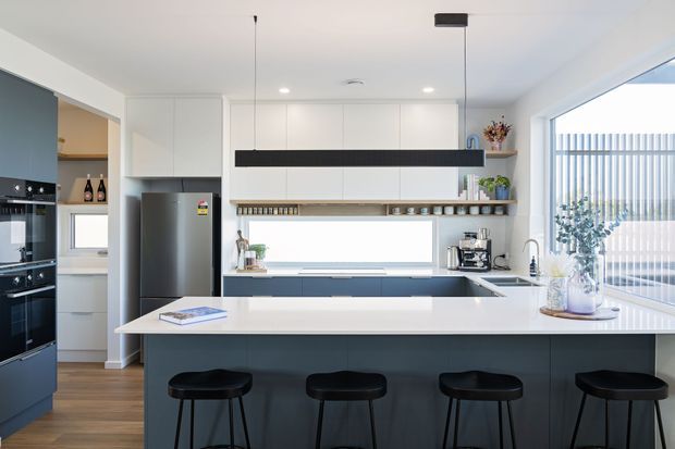 Bright contemporary kitchen with a large island and four black bar stools, white upper cabinetry, dark lower cabinetry, integrated black appliances, timber flooring, open shelving, and broad glazed windows with vertical blinds. A long black linear pendant provides a strong horizontal feature above the island.