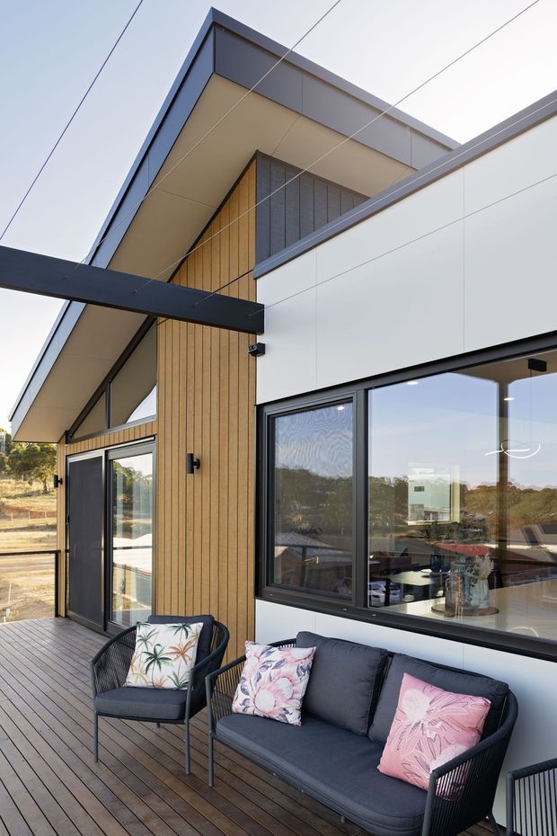 A contemporary residential exterior viewed from a timber-clad deck, featuring a crisp white and dark-grey facade, vertical timber wall cladding, large black-framed windows, and a dramatic projecting roof canopy supported by a dark steel beam. Outdoor lounge furniture occupies the deck, while the interior is visible through the glazing.
