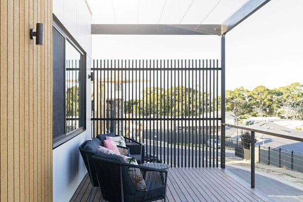 A bright contemporary balcony or terrace with dark timber decking, black metal vertical screening and balustrades, and outdoor lounge chairs beside a glazed and timber-clad building facade. The elevated terrace overlooks neighbouring rooftops and leafy suburban surroundings.