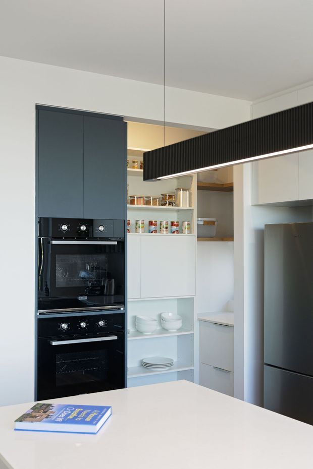 A contemporary kitchen with a tall dark built-in oven tower, white cabinetry, open pantry shelving, integrated refrigerator, and a large white island. A long black ribbed pendant light hangs above the island, adding a strong linear feature.