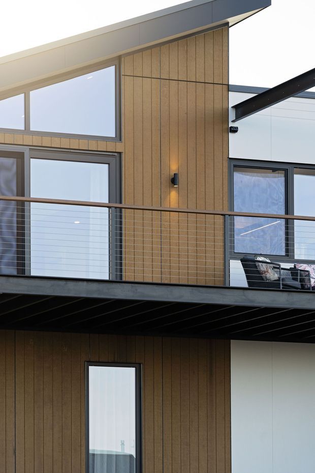 Close-up exterior view of a contemporary multi-level residential building with warm vertical timber-look cladding, crisp white wall panels, dark metal roof and structural elements, large glazed windows, and a projecting balcony with thin horizontal cable railing. A small outdoor seating area is visible on the upper balcony.