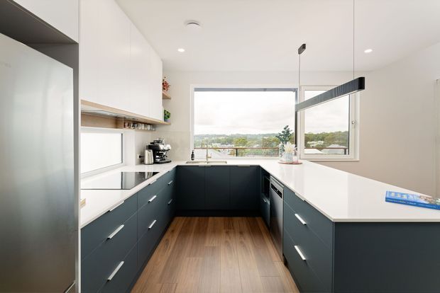 Bright contemporary U-shaped kitchen with dark matte lower cabinetry, white upper cabinets and white benchtops. A timber-look floor adds warmth, while large windows provide views over a leafy suburban landscape. Integrated appliances, open shelving and a linear black pendant light complete the streamlined design.