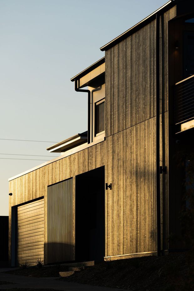 Exterior view of a contemporary multi-level residential building with tall vertical timber cladding, dark metal-framed openings, projecting eaves, and a partially visible garage door. The facade is strongly lit by warm low-angle sunlight against a clear sky.