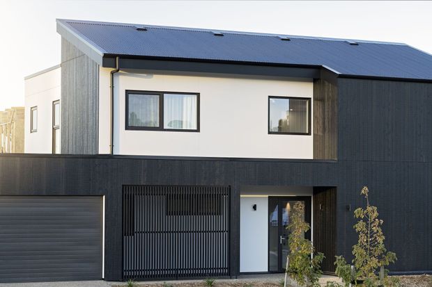 Contemporary two-storey residential facade with a dark corrugated metal gabled roof, black vertical cladding and fencing, white rendered upper walls, black-framed windows, and an integrated garage and recessed entry. The clean geometric composition is viewed from the street, with minimal landscaping in the foreground.