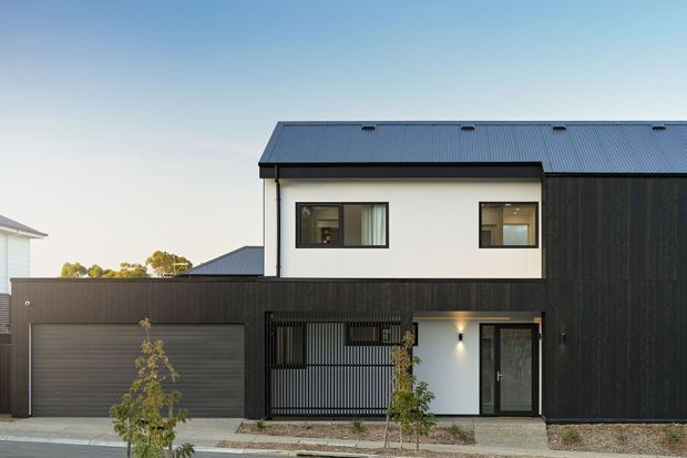 Street-facing view of a contemporary multi-level townhouse with a restrained black, charcoal and white facade. The building has a low-pitched corrugated metal roof, dark vertical cladding, white rendered upper walls, a large integrated garage, screened entry fencing, and a glazed front door. A paved pedestrian frontage and sparse planting complete the modern urban composition.