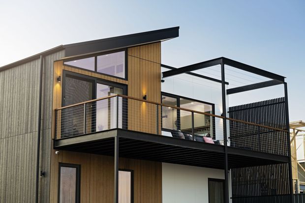Exterior view of a contemporary multi-storey building with vertical timber-look cladding, dark metal framing, a projecting upper balcony with wire balustrade, and a partially covered rooftop or terrace structure. Large glazed openings and clean geometric rooflines create a modern architectural composition.