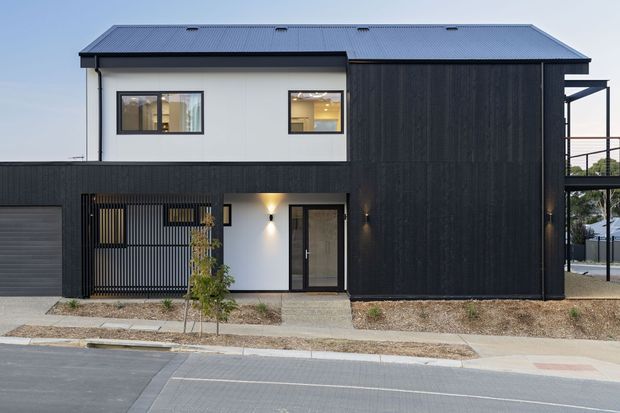 Street-facing view of a contemporary two-storey house with a simple gabled metal roof, crisp white upper-level walls, dark vertical cladding, black-framed windows, an integrated garage, and a covered entry area with slatted screening.